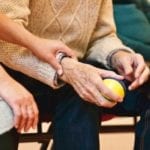 An elderly man sits holding a ball while another person gently grabs his wrist