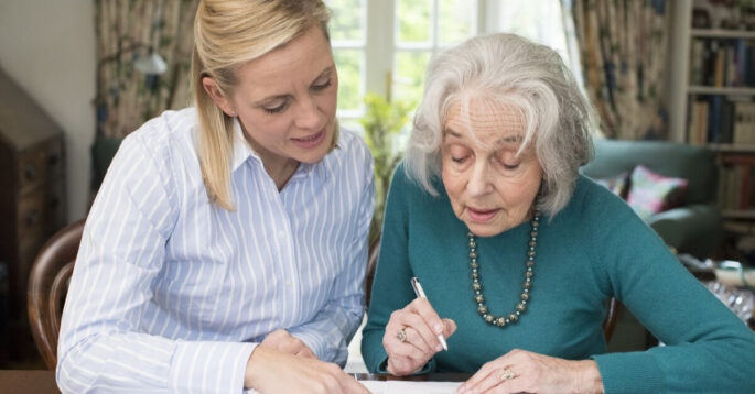 A middle-aged woman helps an elderly woman sign some documents