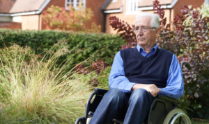 An older man in a wheelchair sits outside a nursing home with a concerned look on his face