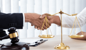 Two people shake hands over a table with a gavel, legal documents, and scales of justice.