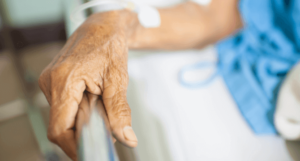 An older adult's hand resting on a hospital bed rail, with an IV line secured with medical tape.