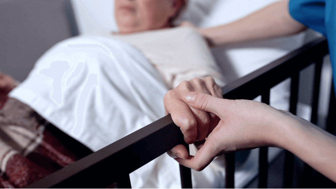 An older person sits in bed while a younger person comforts them