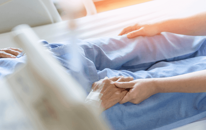 A caregiver gently holds the hand of an older adult in a hospital bed.