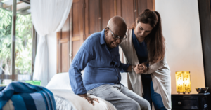 An older man is helped out of bed by a caregiver to prevent nursing home falls.