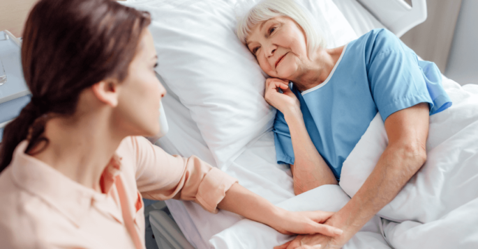 a female caregiver sits next to an older woman who is lying in bed
