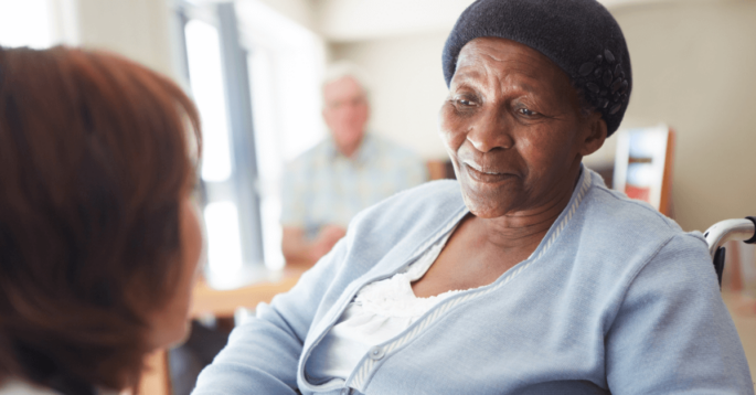 an older woman sits in a nursing home and looks at a nurse in the foreground