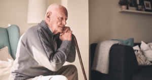 An older man looks worried while sitting in a nursing home, holding a cane.