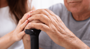A close-up of an older adult's hands resting on a cane, with a caregiver's hands placed gently over them.