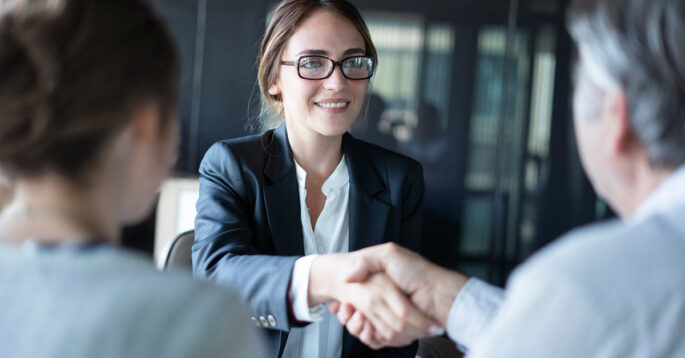 A professional woman with glasses shakes a man's hand across from her, while another woman sits next to him.