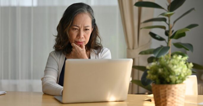 A woman sitting at a table, looking thoughtfully at her laptop screen.