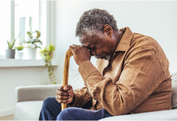 An older adult sits on a couch, leaning forward with his head resting on his hand while holding a cane.