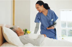 A nurse speaking with an older adult resting in a nursing home bed.