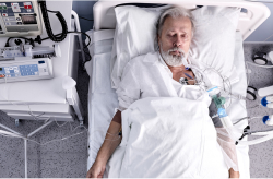 An older man lying unconscious in a hospital bed, connected to medical equipment.