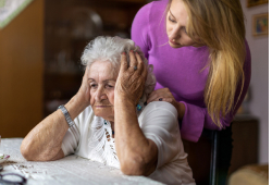 Elderly woman resting her head in her hands, looking distressed, as a younger woman gently leans over her with concern.