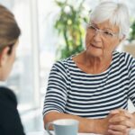 An older woman with white hair and glasses sits at a table speaking seriously with a younger woman, possibly discussing a sensitive issue such as nursing home abuse