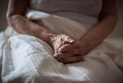 An older adult sits on a bed with their hands clasped in their lap.