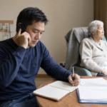 A man talks on the phone and takes notes while an older adult sits by a window in a nursing home room.